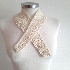 Puede incluir: Bufanda de punto color crema con borde festoneado, exhibida en un maniquí. La bufanda está cruzada en la parte delantera, mostrando su diseño texturizado y detalles delicados. El color neutro la hace versátil.