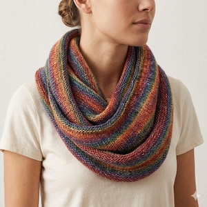Puede incluir: Bufanda infinita tejida en colores del arcoíris, con rayas rojas, naranjas, amarillas, verdes, azules y moradas. La bufanda está envuelta alrededor del cuello de una persona, mostrando el diseño vibrante. La textura suave y los tonos cálidos sugieren un accesorio acogedor.
