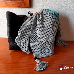 Puede incluir: Un chal de ganchillo gris con un borde turquesa y amarillo y una borla gris. El chal está drapeado sobre un bolso de cuero negro.