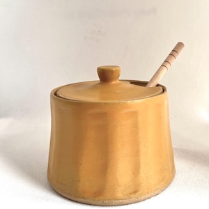 May include: A golden-yellow ceramic sugar bowl with a lid and a small wooden spoon. The spoon is placed inside the bowl. The sugar bowl is a kitchen or dining accessory. The bowl and lid are a warm, golden-yellow colour.