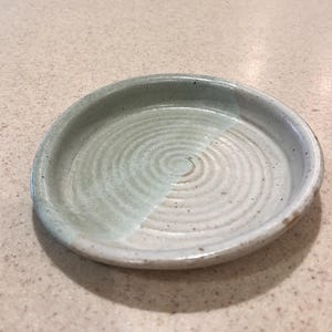 May include: A handmade ceramic dish with a shallow, round design. The dish has a two-tone colour scheme, with a light green and off-white glaze. The interior has a spiral pattern. This pottery piece is suitable for serving or display.