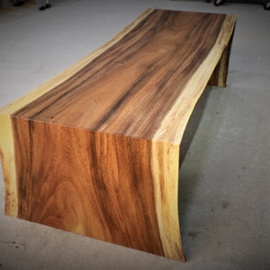 May include: A long, wooden coffee table with a natural, live edge design. The table is made of dark brown wood with lighter brown streaks throughout. The table has a rectangular shape with rounded edges.
