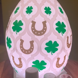 May include: White ceramic diffuser with a shamrock and horseshoe design. Three essential oil bottles are in the foreground: Copaiba, Southern Lights Black Spruce, and Nutmeg.