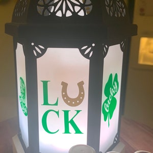 May include: Black lantern with a white frosted glass panel featuring the words "LUCK" with a gold horseshoe in the center. Green shamrocks with the word "lucky" are on either side of the lantern. Three essential oil bottles are in the foreground.