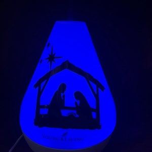 Może przedstawiać: Biały dyfuzor z niebieskim światłem i sylwetką sceny narodzin. Dyfuzor ma napis "Young Living" z przodu.