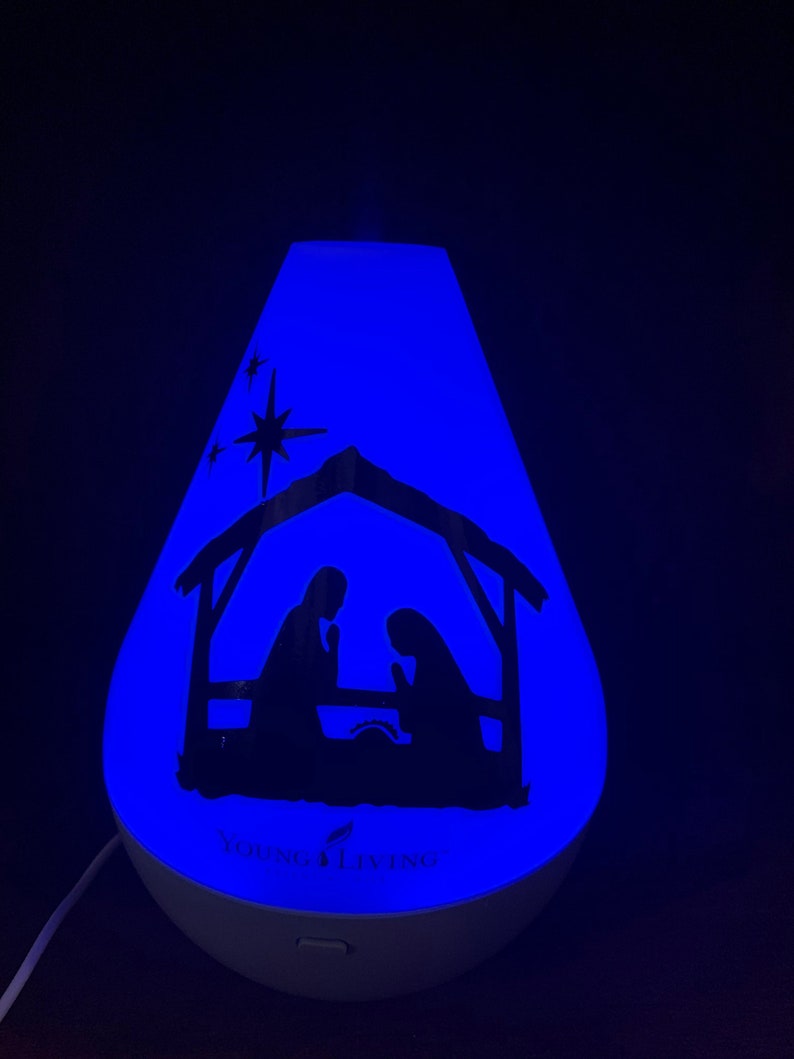 Może przedstawiać: Biały dyfuzor z niebieskim światłem i sylwetką sceny narodzin. Dyfuzor ma napis "Young Living" z przodu.