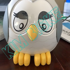 May include: A white owl-shaped piggy bank with yellow feet and black eyes. The owl has a winking expression.