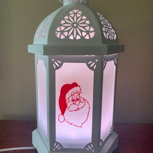 May include: White lantern with a red Santa Claus design on the front. The lantern has a white cord and a white base.