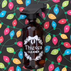 May include: A brown glass spray bottle with a black trigger sprayer. The bottle has a white label with a wreath design and the text "Thieves Cleaner".