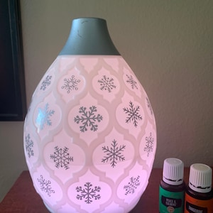 May include: A white ceramic diffuser with a snowflake design and a teal top. The diffuser is lit up and has two bottles of essential oil next to it. The bottles are labeled "Peace & Calming" and "Tangerine".