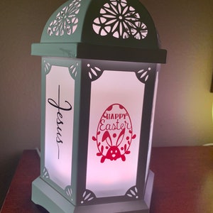May include: A white lantern with a cut-out design on the top and sides. The lantern has a white background with the words "Jesus" and "Happy Easter" in red lettering. The lantern is lit from within, casting a warm glow. The lantern is made by Young Living.