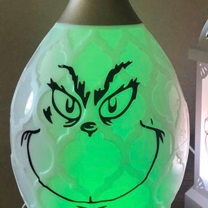 May include: A white diffuser with a green light and a black Grinch face design. The diffuser is labeled "Young Living".