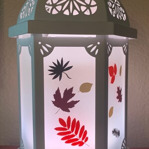 May include: White ceramic lantern with a cut-out design and a frosted glass panel featuring a fall leaf design. The lantern has a base with the text "YOUNG LIVING" printed on it.