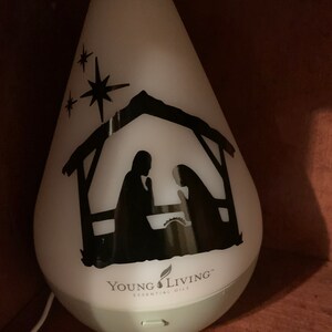 Może przedstawiać: Biały dyfuzor z czarną sylwetką sceny narodzin i trzema gwiazdami. Dyfuzor ma napis "Young Living Essential Oils" na dole.