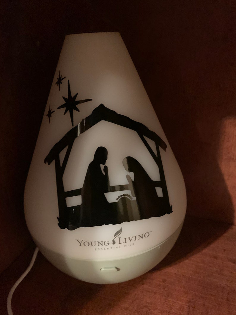 Może przedstawiać: Biały dyfuzor z czarną sylwetką sceny narodzin i trzema gwiazdami. Dyfuzor ma napis "Young Living Essential Oils" na dole.