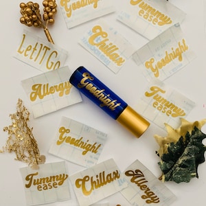May include: A blue glass roller bottle with a gold cap labeled "Goodnight".  The bottle is surrounded by gold and white stickers with various phrases, including "Tummy Ease", "Allergy", "Let It Go", "Chillax", and "Goodnight".