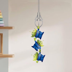 Peut inclure: Trois jouets extraterrestres verts à trois yeux chacun, vêtus d'uniformes bleus et violets, sont suspendus à une pince argentée. Les jouets sont tirés du film Toy Story.