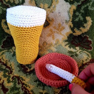 Könnte beinhalten: Ein gehäkeltes Bierglas mit einem weißen Schaum oben und ein gehäkelter Aschenbecher mit einer Zigarette darin. Die Gegenstände befinden sich auf einem gemusterten Teppich.