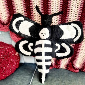 Könnte beinhalten: Eine schwarze und weiße gehäkelte Motte mit einem Skelett-Design. Die Motte sitzt auf einer rosa und rot gestreiften gehäkelten Decke.