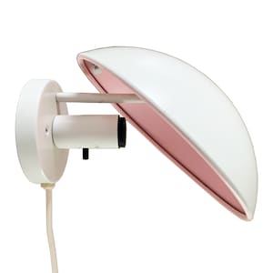 May include: A white wall-mounted lamp with a curved, wing-like shade. The interior of the shade is a soft pink, contrasting with the white exterior. The lamp has a cylindrical base and a cord for power.