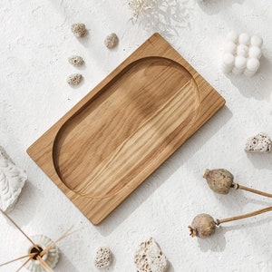 Puede incluir: Una bandeja rectangular de madera con un hueco ovalado redondeado. La bandeja está sobre una superficie blanca con flores secas, piedras blancas y otros elementos decorativos.