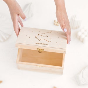 The picture shows two hands partly opening the natural wooden box with an engraved constellation sign. It shows the natural wooden insides of the box.