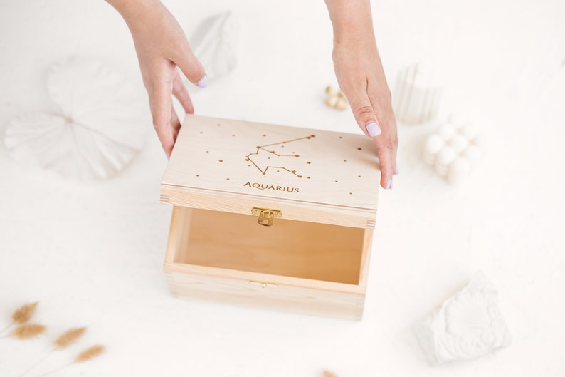 The picture shows two hands partly opening the natural wooden box with an engraved constellation sign. It shows the natural wooden insides of the box.