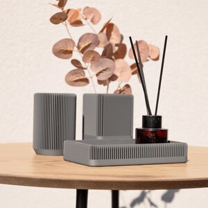 May include: A set of three gray, ribbed ceramic containers with a gray, ribbed tray. The containers are different sizes and are arranged on a wooden table. A glass bottle with black sticks is in the center container.