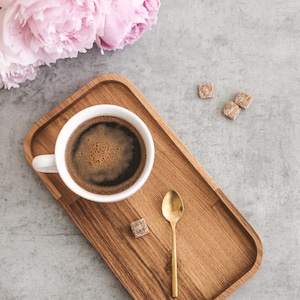 Pode incluir: Uma xícara de café branca com uma colher de ouro e cubos de açúcar em uma bandeja de madeira. Há peônias cor-de-rosa no fundo.