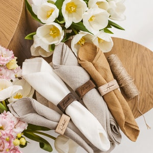 May include: Four linen napkins with wooden napkin rings engraved with the names "Robert", "Nick", "Mary", and "Anna". The napkins are arranged on a wooden surface with a bouquet of white tulips and pink flowers.