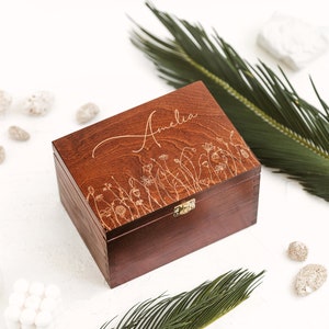 The picture shows redwood box with metal clasp and engraving of a meadow and a name Amelia, which can be personalized. The box is positioned on the white background with two palm leaves and white stones.