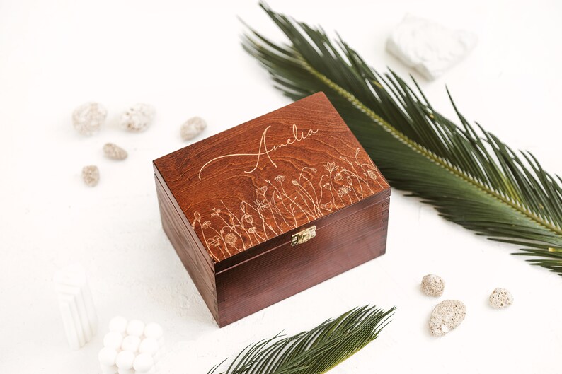 The picture shows redwood box with metal clasp and engraving of a meadow and a name Amelia, which can be personalized. The box is positioned on the white background with two palm leaves and white stones.