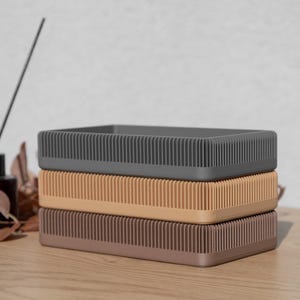 Puede incluir: Tres contenedores de almacenamiento rectangulares apilados uno encima del otro. El contenedor superior es gris, el contenedor del medio es beige y el contenedor inferior es marrón.