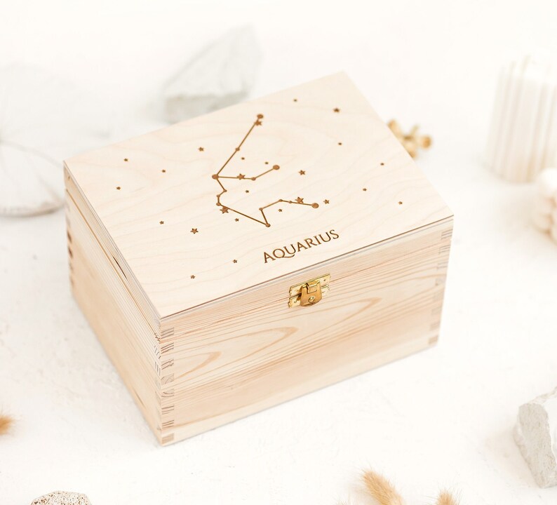 Box made out of natural wood shown sideways. The top of the box has an engraving of the Aquarius constellation sign and small stars. Wooden box comes with a metal clasp.