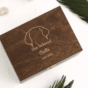 Dark brown wooden box featuring an engraving of a silhouette of golden retriever dog ears and the word: Our beloved Bella and the dats 2009-2023. The ears, the name and the dates can be personalized in the personalization section.