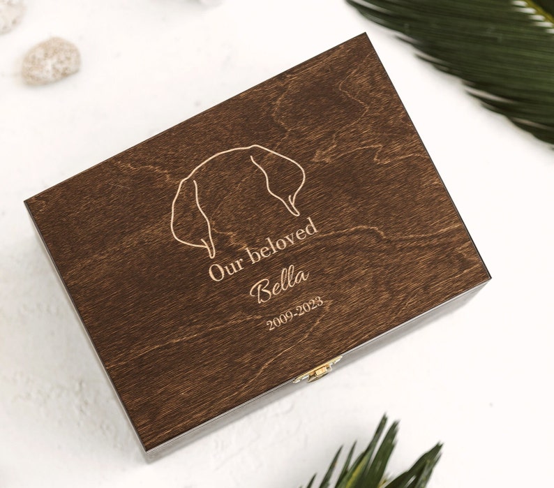 Dark brown wooden box featuring an engraving of a silhouette of golden retriever dog ears and the word: Our beloved Bella and the dats 2009-2023. The ears, the name and the dates can be personalized in the personalization section.