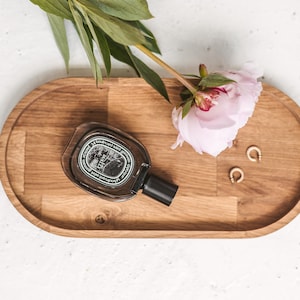 Puede incluir: Una bandeja ovalada de madera con un acabado marrón oscuro. La bandeja contiene un frasco de perfume, una peonía rosa y dos pendientes de aro dorados.