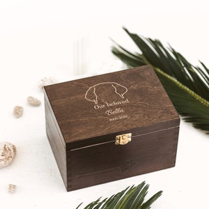 Dark brown wooden pet grief box with golden clasp shown sideways. Surrounded with two palms leaves and various stones. The box is used to store toys and other items of the deceased dog to keep as the cherished memories which were made.