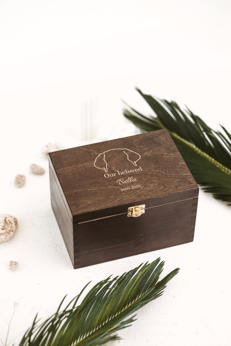 Dark brown wooden pet grief box with golden clasp shown sideways. Surrounded with two palms leaves and various stones. The box is used to store toys and other items of the deceased dog to keep as the cherished memories which were made.