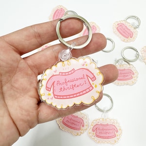 May include: Pink acrylic keychain with a cloud-shaped design featuring a pink sweater with the text "Professional thrifter!" in white. The keychain is attached to a silver key ring.