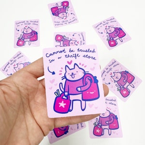 May include: A set of pink and purple stickers with a cartoon cat holding shopping bags. The text on the stickers reads "Cannot be trusted in a thrift store".