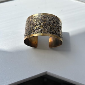 May include: A wide brass cuff bracelet featuring an intricate, dark-etched botanical pattern of intertwined leaves and vines across the outer surface. The metal has a polished gold-tone finish with a visible gap at the back for an open design.