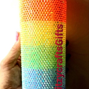 May include: A rainbow-colored tumbler covered in iridescent rhinestones. The tumbler is held in a hand and the text "@glitzycraftsgifts" is visible on the side.