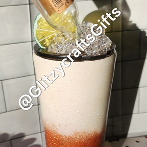 May include: A white and copper glitter tumbler with a clear glass bottle, a lime wedge, and ice cubes inside. The bottle has a brown label with the text "Titos Vodka.com 80 Proof 50 mL".