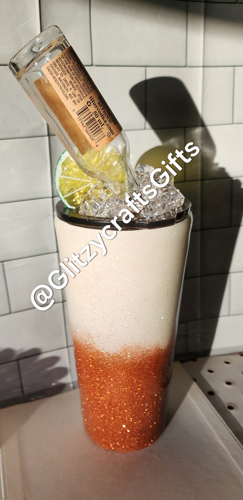 May include: A white and copper glitter tumbler with a clear glass bottle, a lime wedge, and ice cubes inside. The bottle has a brown label with the text "Titos Vodka.com 80 Proof 50 mL".