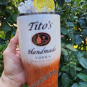 May include: A white and orange glitter tumbler with the words "Tito's Handmade Vodka Austin Texas" and a logo. The tumbler has a clear lid with a lemon slice and ice cubes.