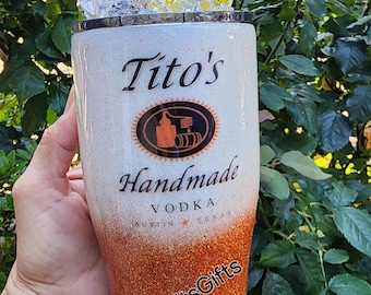 Könnte beinhalten: Ein weißer und orangefarbener Glitzerbecher mit den Worten "Tito's Handmade Vodka Austin Texas" und einem Logo. Der Becher hat einen durchsichtigen Deckel mit einer Zitronenscheibe und Eiswürfeln.
