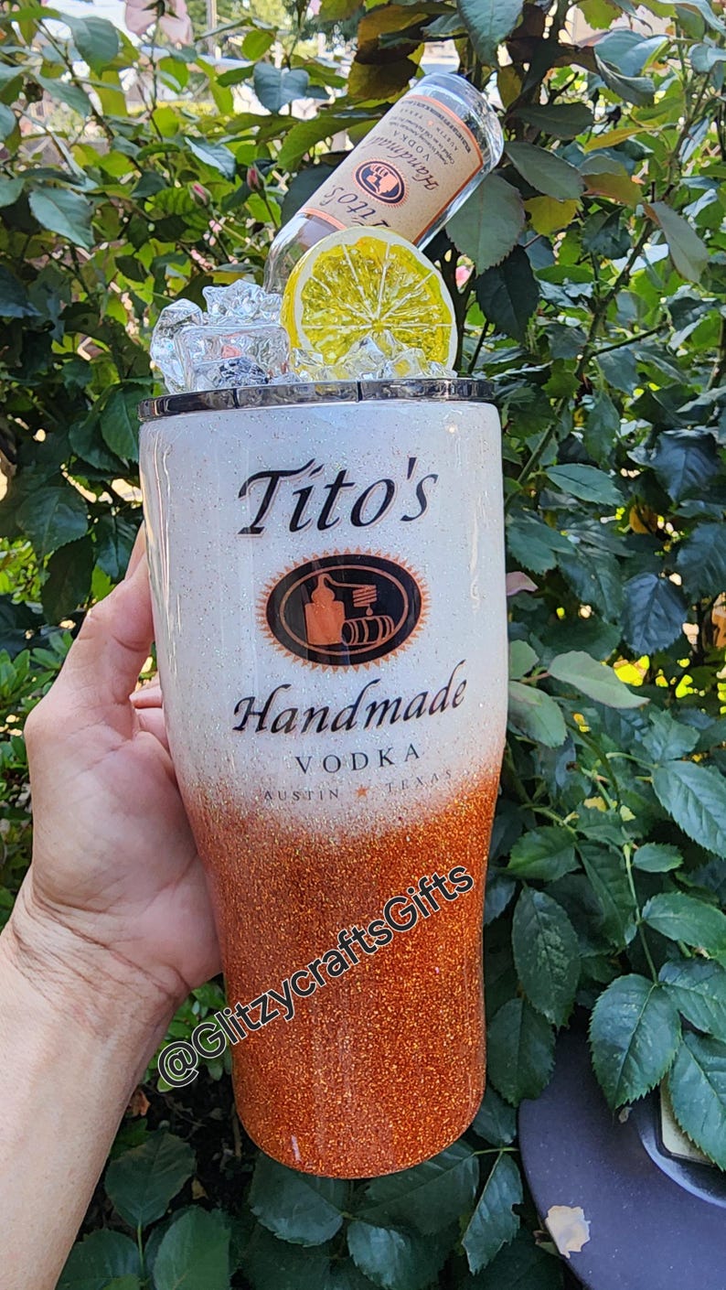 May include: A white and orange glitter tumbler with the words "Tito's Handmade Vodka Austin Texas" and a logo. The tumbler has a clear lid with a lemon slice and ice cubes.