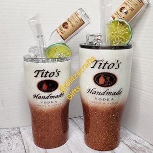 May include: Two white and copper glitter tumblers with a Tito's Handmade Vodka logo. The tumblers are filled with ice and a lime wedge. The tumblers are on a white wood background.