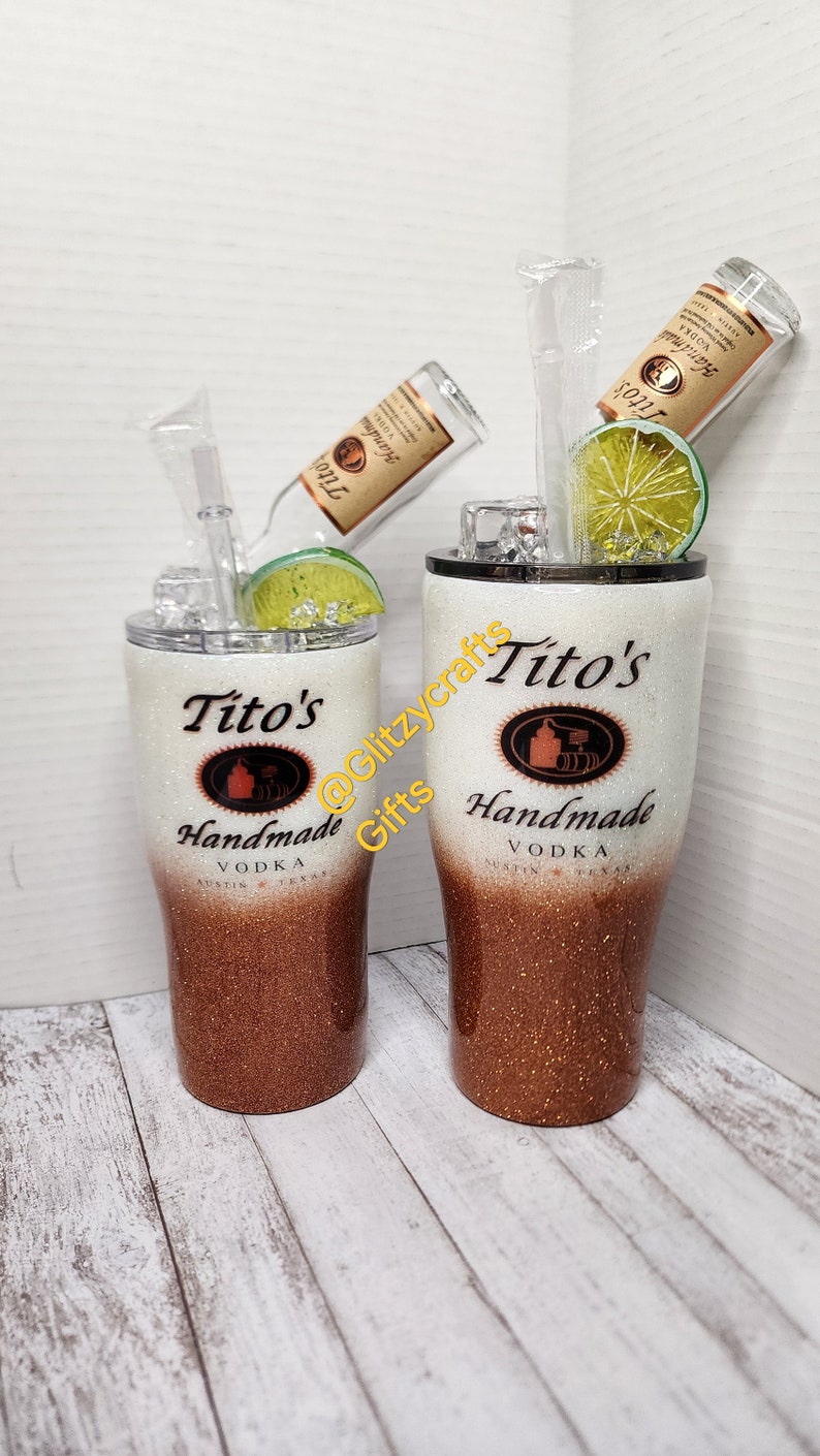 May include: Two white and copper glitter tumblers with a Tito's Handmade Vodka logo. The tumblers are filled with ice and a lime wedge. The tumblers are on a white wood background.